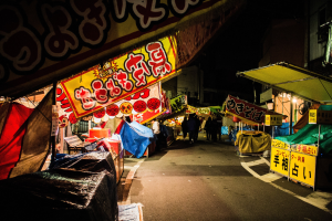 Ein belebter Nachtmarkt mit zahlreichen beleuchteten Ständen, die eine Straße säumen, Menschen, die zwischen ihnen hindurchgehen, und Gebäude auf beiden Seiten, mit einer Person im Vordergrund.