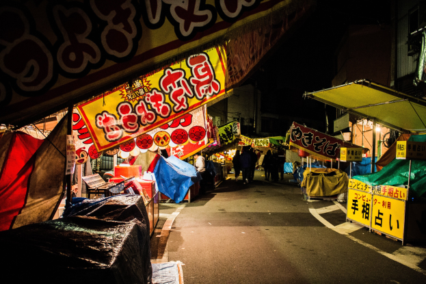 Ein belebter Nachtmarkt mit zahlreichen beleuchteten Ständen, die eine Straße säumen, Menschen, die zwischen ihnen hindurchgehen, und Gebäude auf beiden Seiten, mit einer Person im Vordergrund.