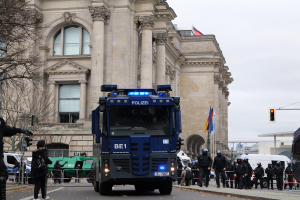 Eine Gruppe von Polizisten steht vor einem großen Gebäude mit Fenstern, Säulen und Bögen, mit Fahrzeugen auf der Straße, einer Person mit einer Kamera links und einem klaren blauen Himmel im Hintergrund.