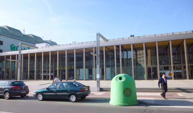 Grüner Müllcontainer auf einer Stadtstraße neben einem Glaswandgebäude mit Säulen, mit Fahrzeugen, Passanten und einem bewölkten Himmel im Hintergrund.