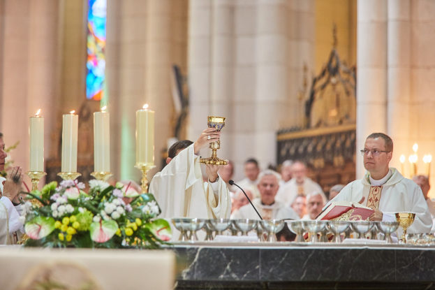 Ein Priester hält einen Kelch vor einer Gruppe von Menschen in einer Kirche, mit Gläsern, einem Mikrofon, einem Buch und einem Blumenstrauß auf einem Tisch im Vordergrund und Kerzen, Säulen und Buntglasfenstern im Hintergrund.