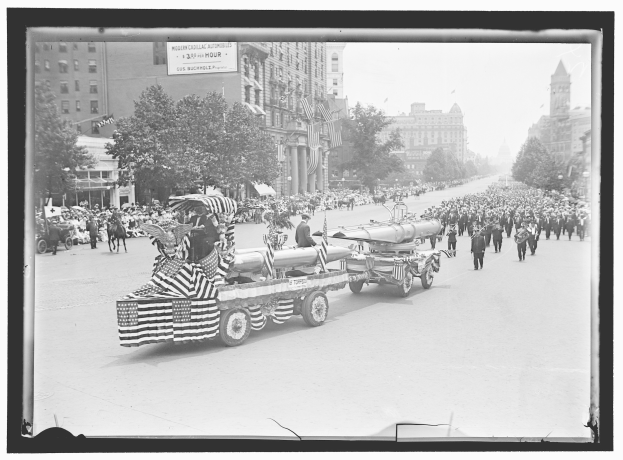 Schwarzes und weißes Foto eines Umzugs mit einem Feuerwehrauto in der Mitte, umgeben von Menschen, einige halten Fahnen, mit Bäumen, Gebäuden und einer Tafel mit Text im Hintergrund.