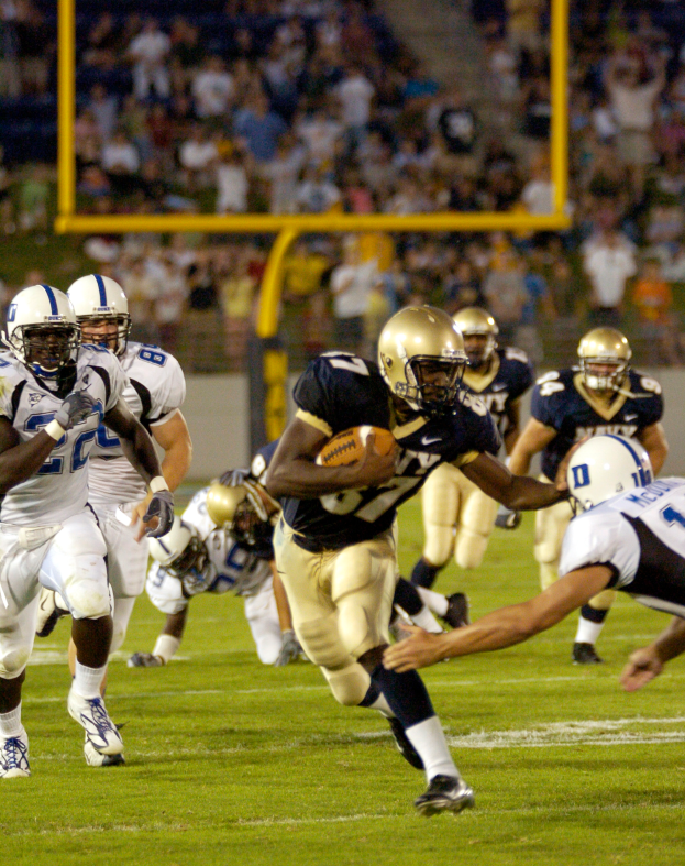 Ein Fußballspieler in einem Sportdress und Helm rennt mit dem Ball, umgeben von anderen Spielern, mit unscharfen Hintergrundelementen und Text am unteren Bildrand.